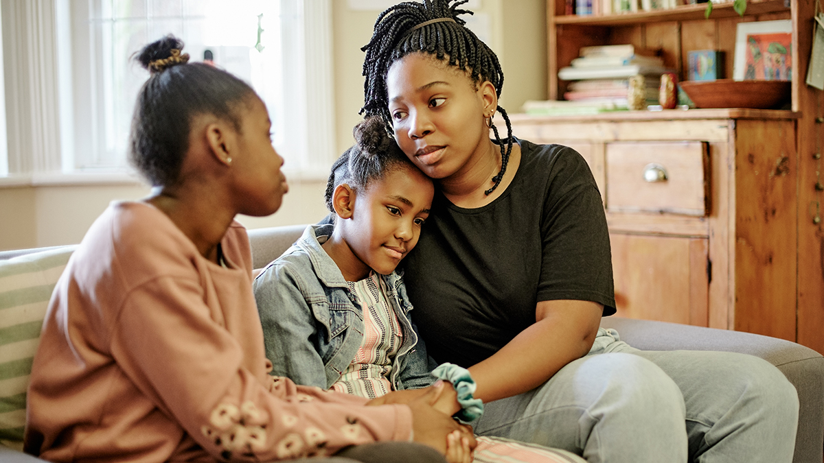 Young mother comforts her two young daughters while seated on a couch.