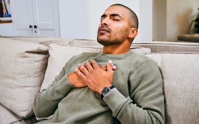A Black person with dark facial hair on the couch with their eyes closed and their hands on their chest.
