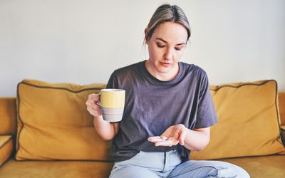 A person seated on a sofa holding a cup in one hand and tablets in the other