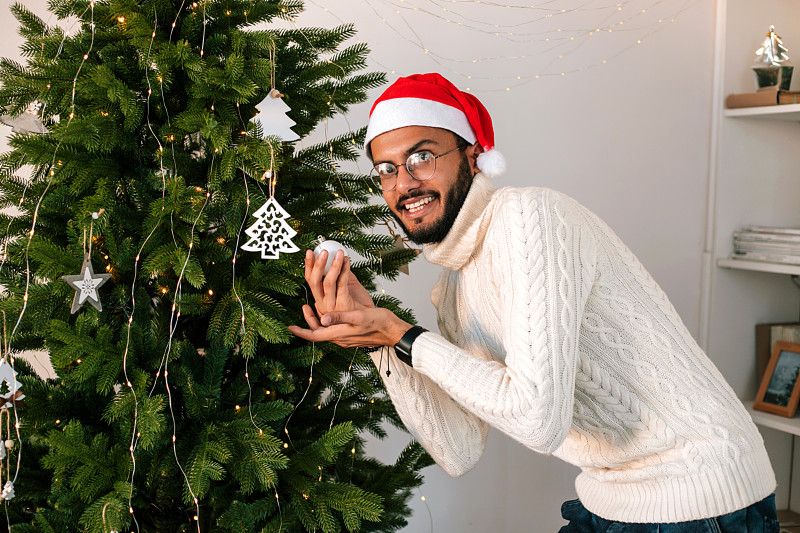 年轻的印度男子在家里装饰圣诞树的画像。图片下载