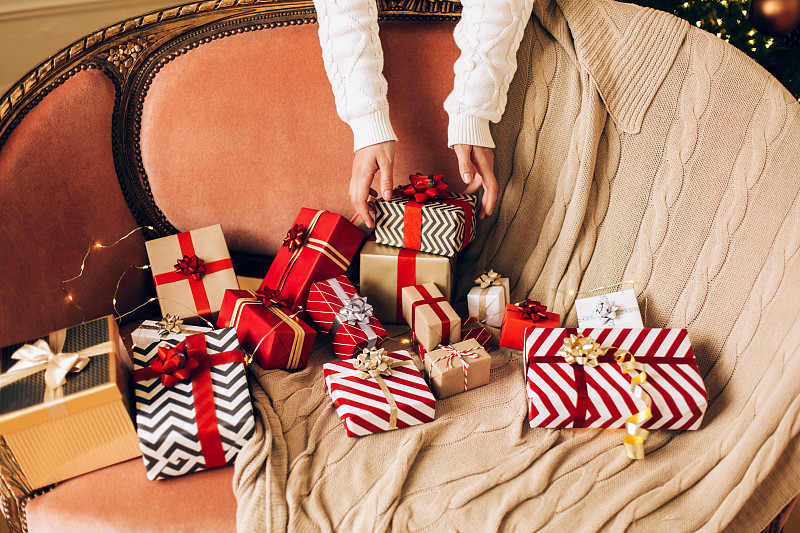 穿着白色针织毛衣的女人正拉着手拿着许多不同的礼品盒，礼品盒上有蝴蝶结，放在有格子和细绳的沙发上。新年庆祝概念。前视图。特写镜头图片下载